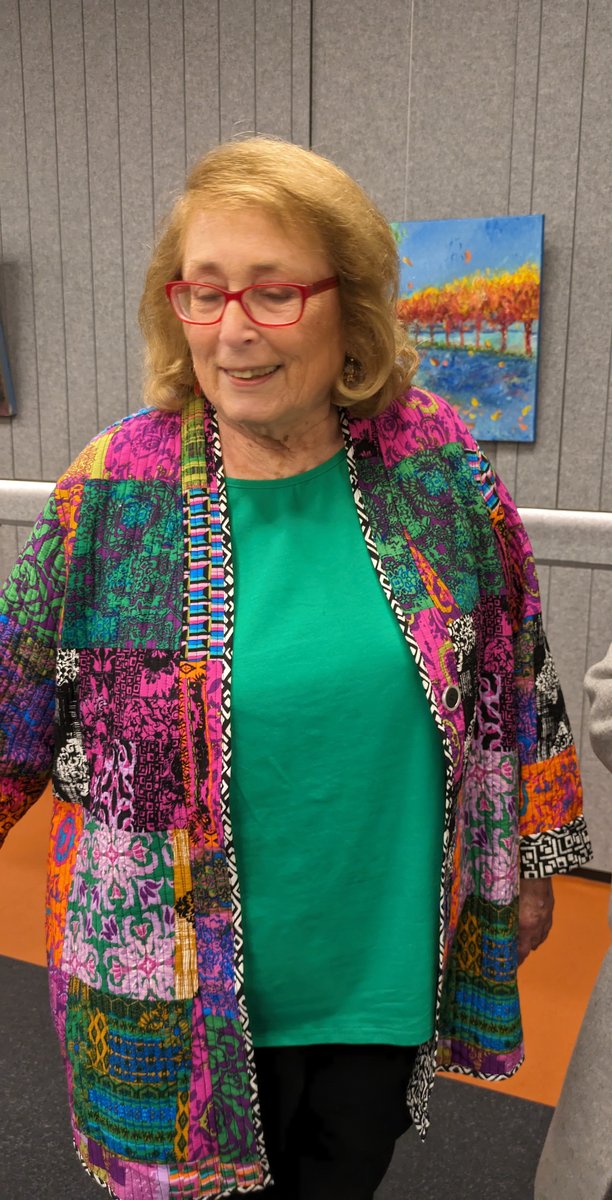 Madeleine Popper smiling in a bright patchwork jacket and red glasses at an exhibition, with her painting Red and Dark on the wall behind her.