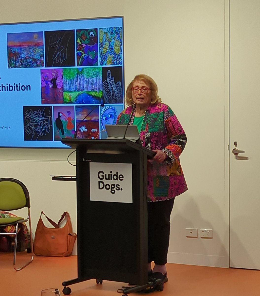 Madeleine Popper speaking at a Guide Dogs lectern at the opening of the Our Vision Works exhibition, with a screen of her artworks behind her.